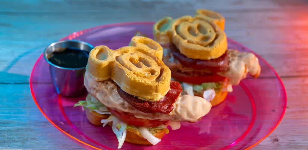 Two Mickey Mouse-shaped sandwiches sit on a pink plate, each filled with crispy lettuce, sliced tomatoes, sauce, and savory sausage. A small metal cup with a dark dipping sauce accompanies the dish. The food is presented on a wooden table, with a playful ambiance created by soft blue and red lighting, highlighting the fun and whimsical theme of the meal.