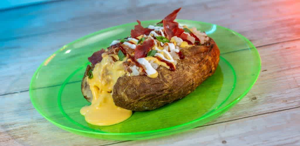 A baked potato loaded with creamy cheese is showcased on a vibrant green plate. Crispy bacon pieces, tangy sour cream, and fresh chives top the potato, which rests on a light wooden surface. The cheese oozes deliciously from the side, creating an appetizing and colorful presentation perfect for that satisfying meal craving.