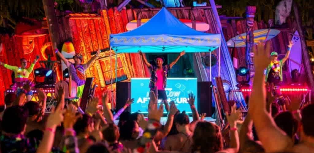 A lively outdoor night party scene with a DJ performing on stage under a blue canopy. The background is decorated with vibrant tropical elements like surfboards and tiki statues. A crowd of people raised their hands, enjoying the energetic atmosphere. Colorful lights illuminate the area, adding to the festive mood.