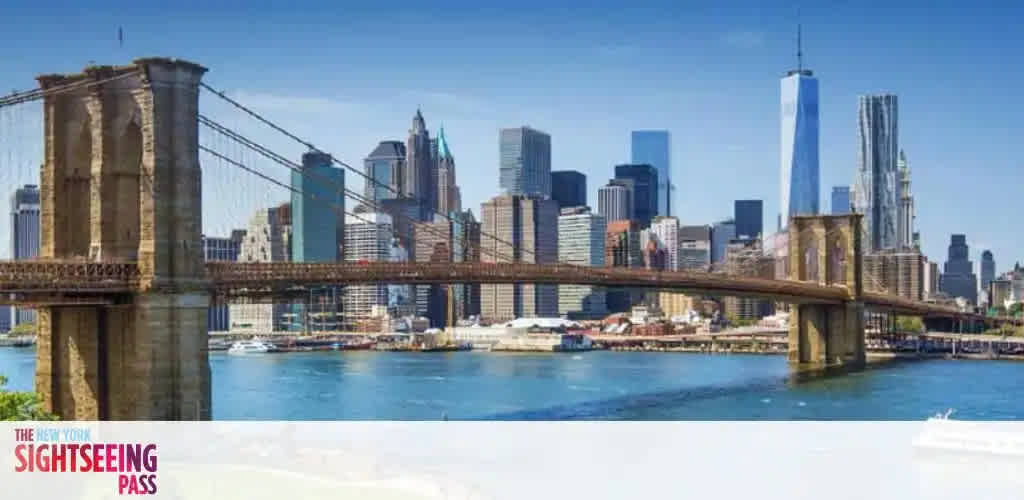 A scenic view of the Brooklyn Bridge in New York City, stretching across the East River. The iconic skyscrapers of Manhattan, including the tallest structure, rise in the background against a clear blue sky. The calm river reflects the bridge and buildings, and a boat is visible. The lower left corner features a logo for The New York Sightseeing Pass.