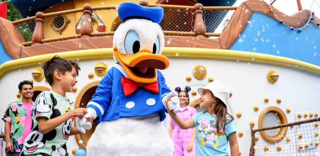 A joyful character in a sailor outfit, with a blue jacket and red bow tie, interacts with two smiling children. They are in front of a colorful nautical-themed backdrop with bubbles in the air. The scene conveys excitement and happiness, perfect for a family-friendly adventure at a theme park. Other children and adults are visible, enjoying the playful atmosphere.