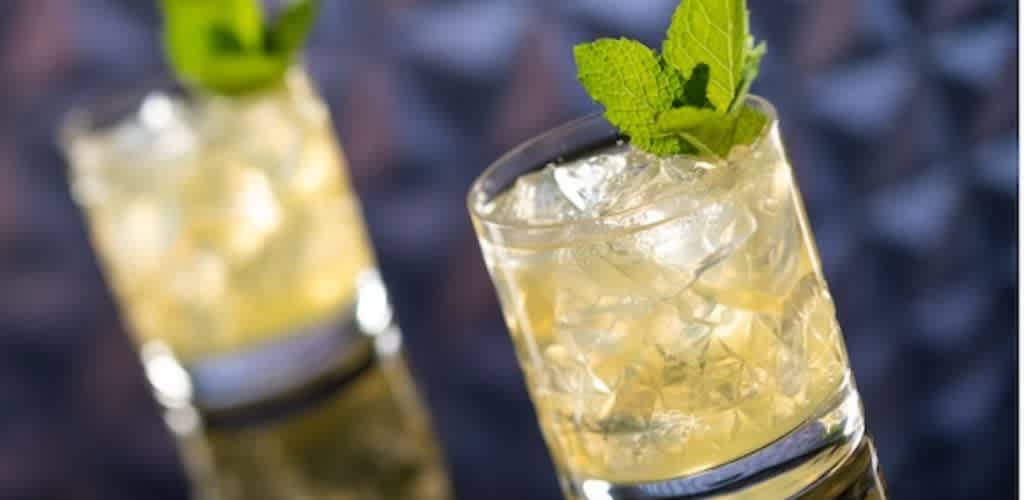Two clear glasses filled with a yellowish beverage over ice are shown. Each glass is garnished with fresh mint leaves. The image is vibrant and the focus is on the front glass, with the second one blurred in the background. The setting appears glossy, creating a reflective surface under the glasses. The overall mood is refreshing and inviting, suggesting a cool, tasty beverage option.