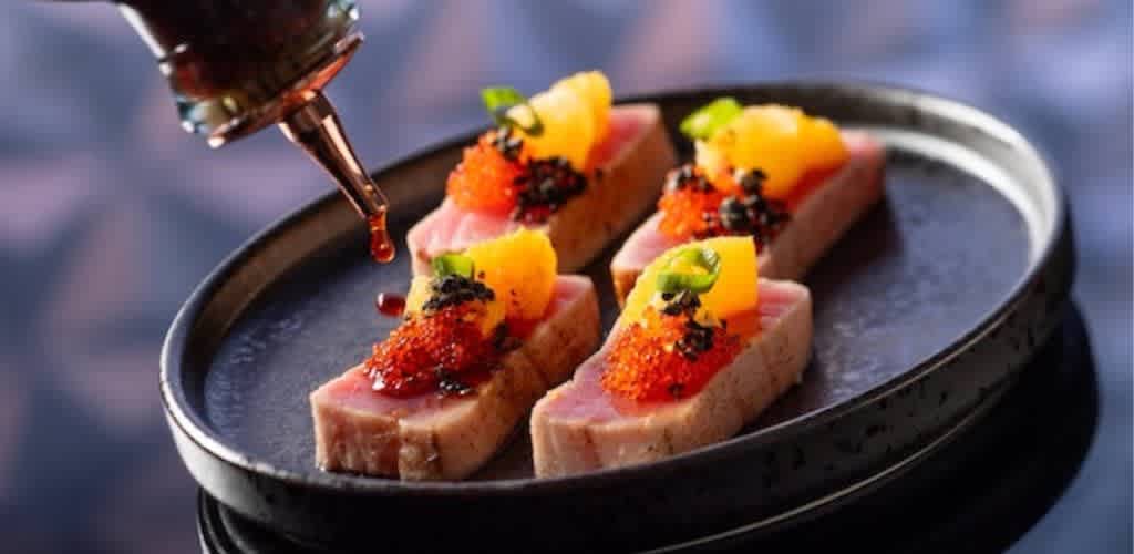 A sleek oval plate holds five pieces of seared tuna topped with vibrant garnishes like orange segments, red roe, and green herbs. A sauce is being artistically drizzled over the tuna, enhancing its appeal. The dish is elegantly presented against a subtle, blurred background, highlighting the rich textures and colors of the ingredients, perfect for a gourmet dining experience.