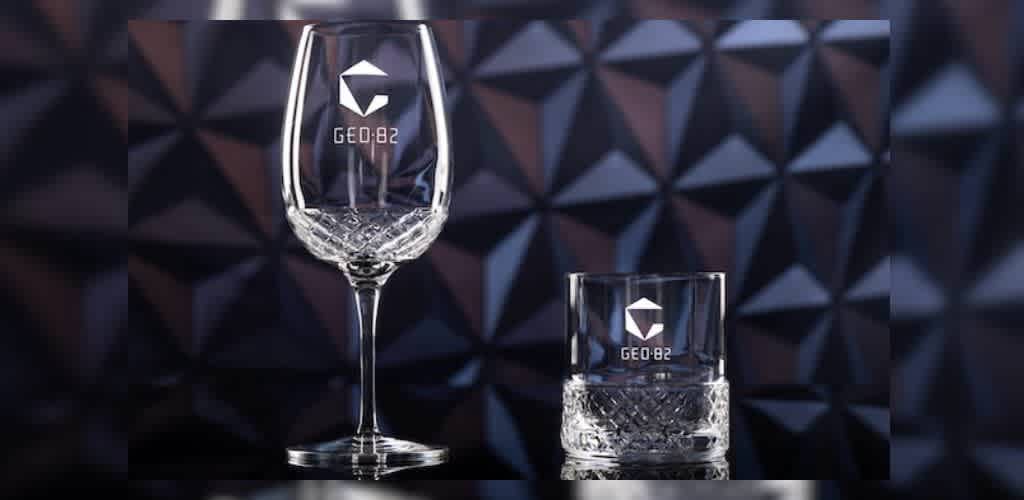 Elegant glassware set displayed against a geometric pattern background. The set includes a tall wine glass and a short tumbler, both featuring intricate cut designs at their bases. The engraving on each piece showcases a geometric logo and the text GEO 82, adding a modern touch. Perfect for enhancing any dining experience.