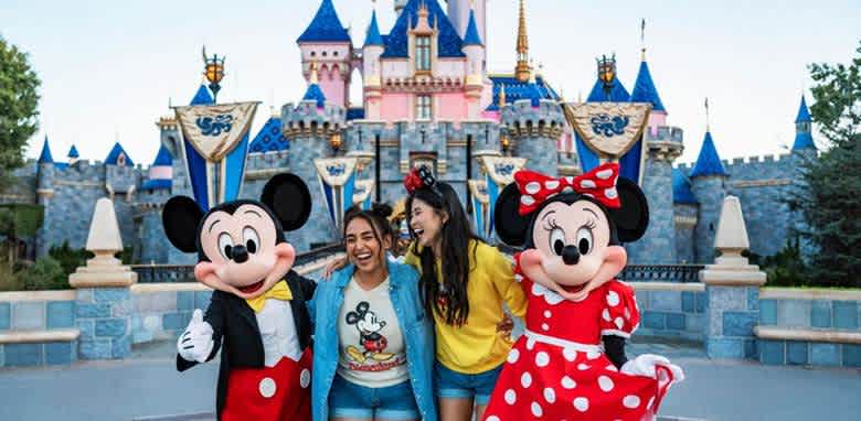 Two people joyfully pose with iconic cartoon characters wearing red and yellow outfits, in front of a dreamy blue and pink castle. The scene is lively and colorful, capturing the excitement of a fun-filled day at a theme park. The characters have large round ears and big smiles, adding to the cheerful atmosphere. The castle in the background is adorned with blue spires and decorative banners, enhancing the magical setting.