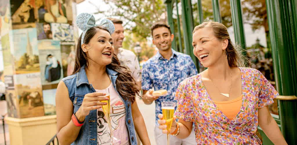A group of four friends enjoy a day at a theme park. Two women in colorful outfits walk in the foreground, each holding a drink and smiling. The woman on the left wears mouse ears, while the woman on the right laughs joyfully. Two men are behind them, one holding a snack. The background shows a green gate and posters, suggesting a lively atmosphere filled with fun and entertainment. The scene conveys joy and relaxation on a sunny day out.