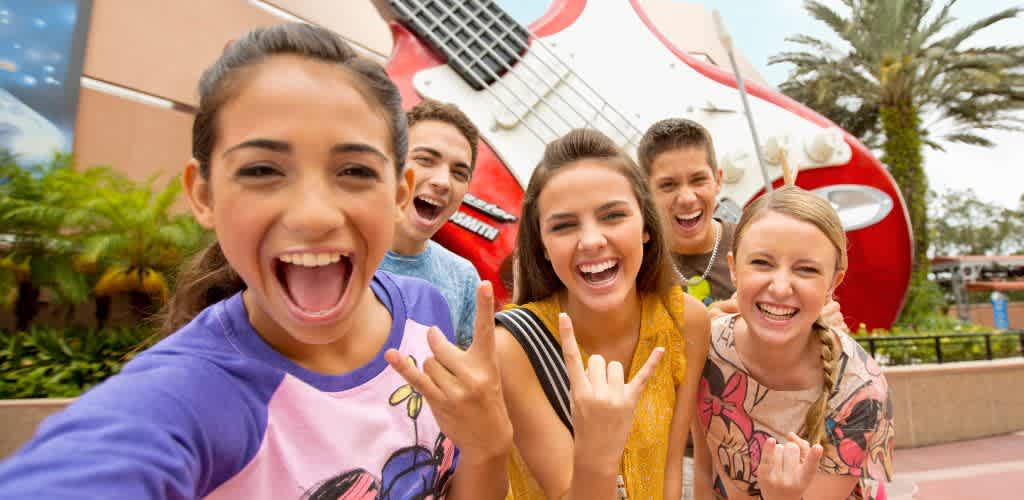 A group of five young adults are joyfully posing in front of a giant red and white electric guitar sculpture. They are making rock and roll hand gestures and smiling widely, enjoying their time at an amusement park. Palm trees and a blue sky are visible in the background, adding to the lively and fun atmosphere.
