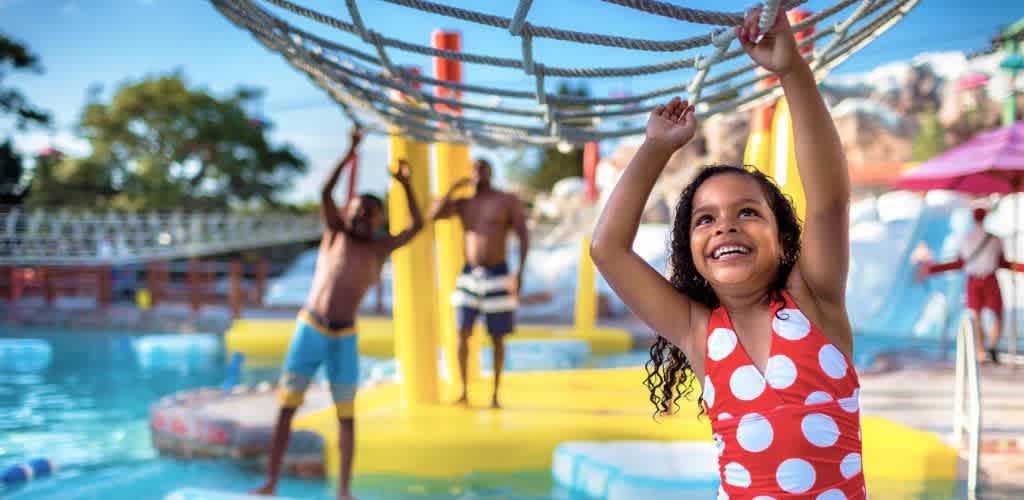 A young girl in a red polka dot swimsuit smiles while playing on monkey bars at a waterpark. Two adults are in the background standing in shallow water. The scene is bright and sunny, with various waterpark structures visible, creating a fun and lively atmosphere for all ages.