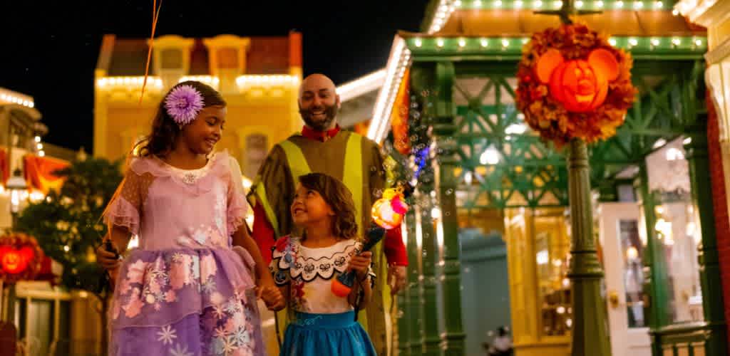 A joyful nighttime scene at a theme park features two children in princess costumes holding glowing wands, walking hand in hand with an adult. They are surrounded by festive lighting and colorful buildings, with a pumpkin-themed decoration on a lamp post. The atmosphere is cheerful and magical, capturing the excitement of a fun-filled Halloween event.