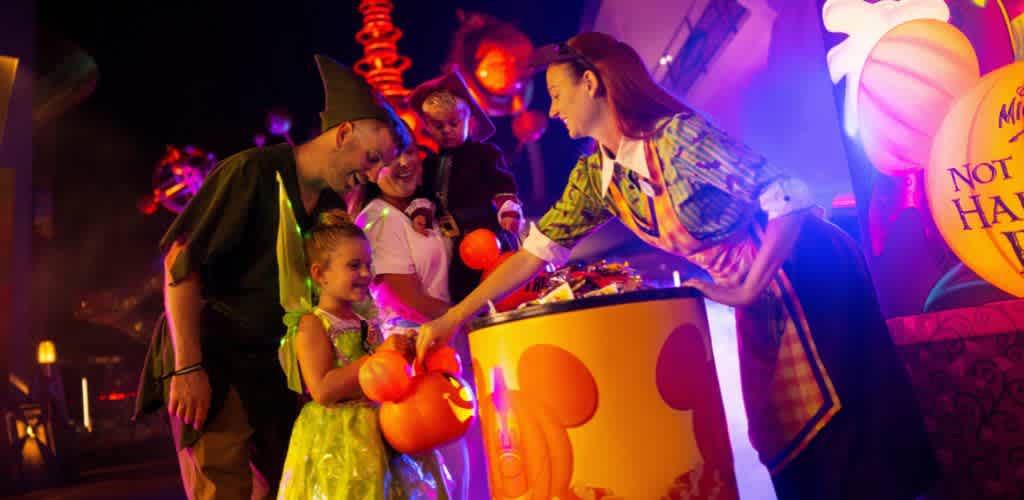 A young child in a green costume receives candy from a person in a colorful outfit. Two adults look on, smiling. The setting is festive with bright decorations and ambient lighting, creating a warm and joyful atmosphere. Orange pumpkins and whimsical elements enhance the playful theme, suggesting a Halloween or party event.
