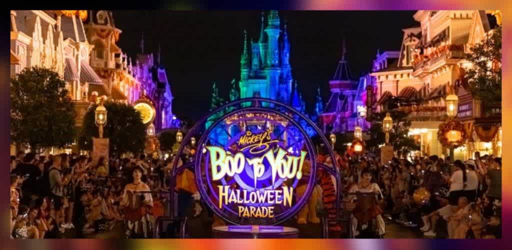 The image shows a vibrant night scene of a Halloween parade at a theme park. A colorful sign in the center reads Boo to You Halloween Parade, with a beautifully lit castle in the background. The street is lined with decorated buildings and filled with a crowd of enthusiastic visitors enjoying the festive atmosphere.