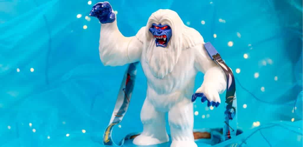 A toy yeti figure stands on a blue backdrop, exuding a sense of adventure. The yeti features a white furred body with a fierce expression and bright blue face and hands. It holds an arm up, revealing sharp teeth, and is adorned with ribbon-like accessories. The background is dotted with small glowing lights, enhancing the whimsical and playful atmosphere. Ideal for festive or imaginative settings.