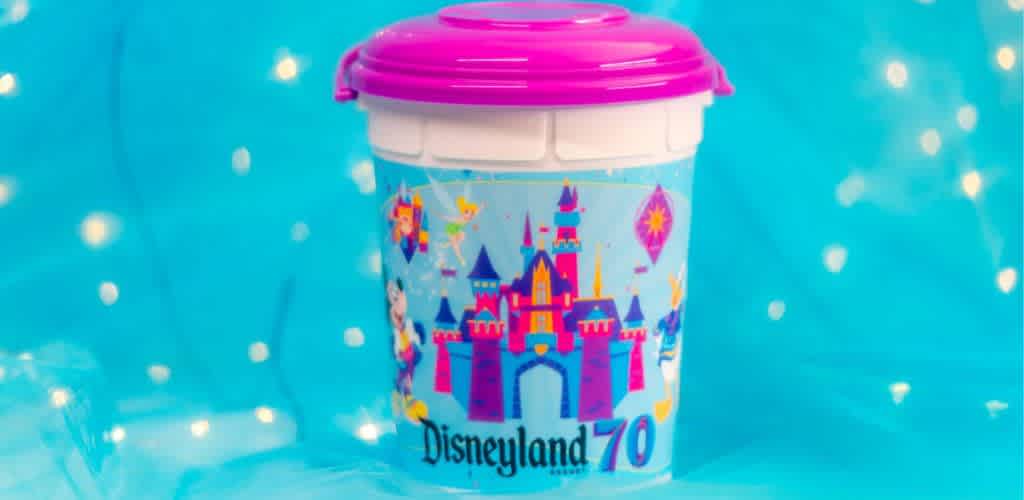 Colorful souvenir cup featuring a vibrant illustration of a castle with playful characters against a bright blue background. The top of the cup is pink, and Disneyland 70 is written on the front. The backdrop is a blurred blue with soft white lights. Perfect for collectors and Disney fans alike, adding a touch of magic to your drinkware collection.