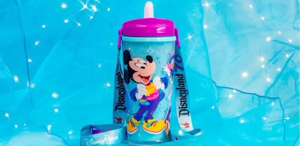 A colorful tumbler features an illustration of a famous cartoon mouse dressed in vibrant clothes, surrounded by playful designs. The tumbler has a pink lid and a built-in straw. A detachable strap with bold text and small graphics enhances its portability. The background is a bright blue with sparkling star-like accents, creating a whimsical atmosphere. Perfect for adding fun to daily hydration.

