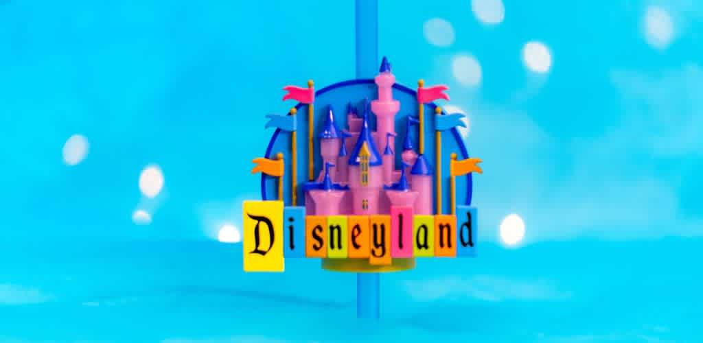 Colorful castle illustration with pink towers and vibrant flags sits on a bright blue background. Below, bold yellow and black letters spell Disneyland. Bokeh light effects add a whimsical touch, evoking a sense of magic and adventure.