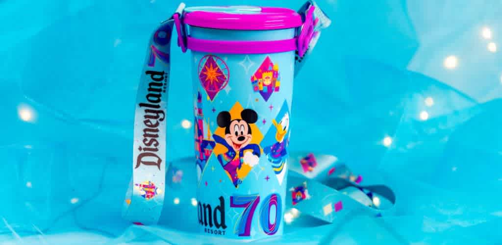 A colorful commemorative Disneyland Resort cup celebrating a 70th anniversary. The design features cartoons against a light blue background, with a vibrant pink lid and matching handle. A lanyard with the word Disneyland in bold lettering is attached. The cup is set on an illuminated blue backdrop that adds a festive, magical feel.