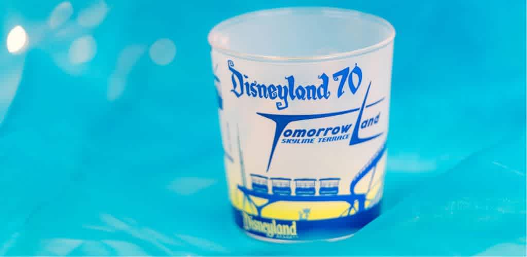 A small, white plastic cup with retro-style blue and yellow graphics featuring the words Disneyland 70 and Tomorrow Land Skyline Terrace, along with an illustration of a futuristic skyline, sits on a turquoise background. The cup evokes a sense of nostalgia and celebration. The background is blurred, emphasizing the cup as the focal point.