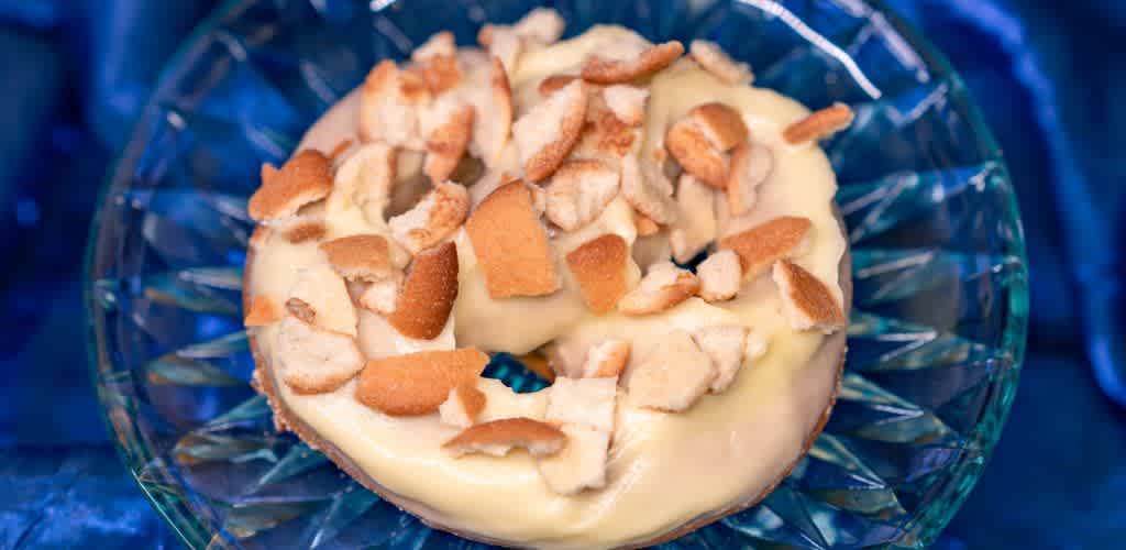 A donut covered in creamy icing and topped with pieces of crumbled cookies sits on an elegant glass plate. The background is a rich blue fabric, enhancing the dessert's presentation.