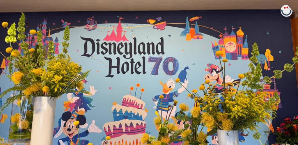 A colorful wall celebrates Disneyland Hotel's 70th anniversary. It features cartoon characters, a pink castle, birthday cakes, and lively patterns. In front, vibrant yellow and green floral arrangements add a cheerful touch, creating a festive atmosphere.
