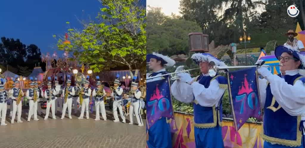 A lively parade scene featuring a marching band in blue and white uniforms playing brass instruments. The band stands in a paved area with vibrant flags displaying the number 70. Trees and colorful decorations create a festive backdrop, capturing the spirit of celebration against a clear evening sky.