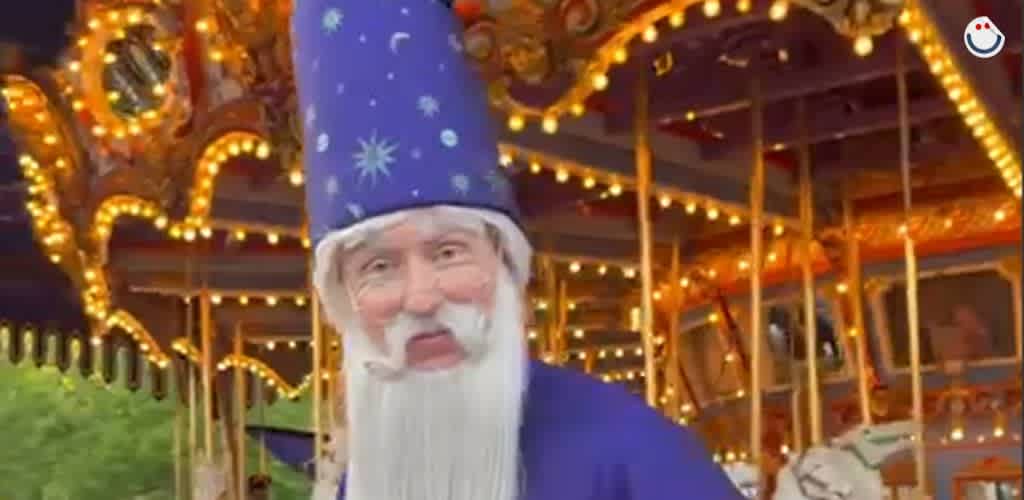 A festive scene with a person dressed as a wizard, wearing a tall blue hat and robe, with a long white beard. They stand in front of a brightly lit carousel adorned with warm, glowing lights. The carousel features decorative details and painted horses, creating a magical, enchanting atmosphere.