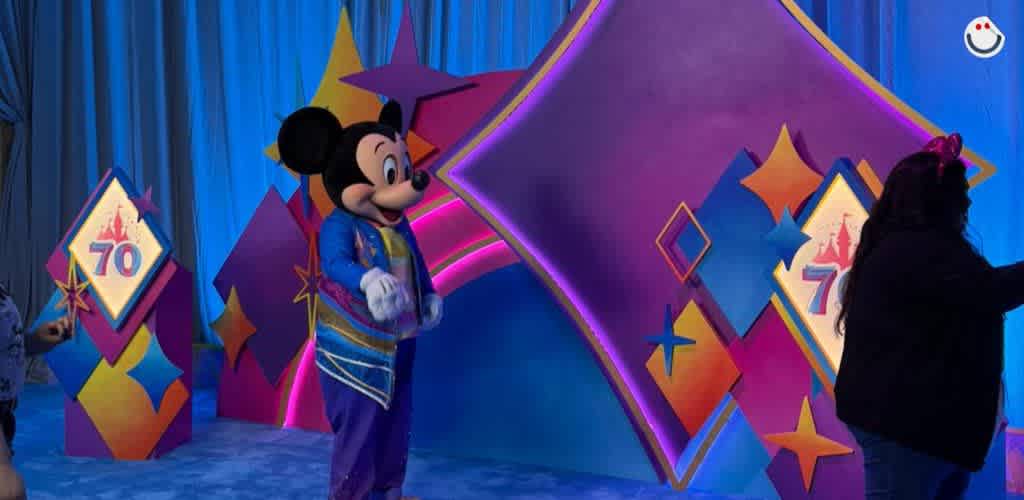 A colorful celebration scene features a costumed character resembling a famous mouse in a blue and purple outfit, posed in front of a vibrant, illuminated geometric backdrop. The setting includes decorative elements that display the number 70, indicating a milestone or anniversary. People are interacting and enjoying the festive atmosphere.