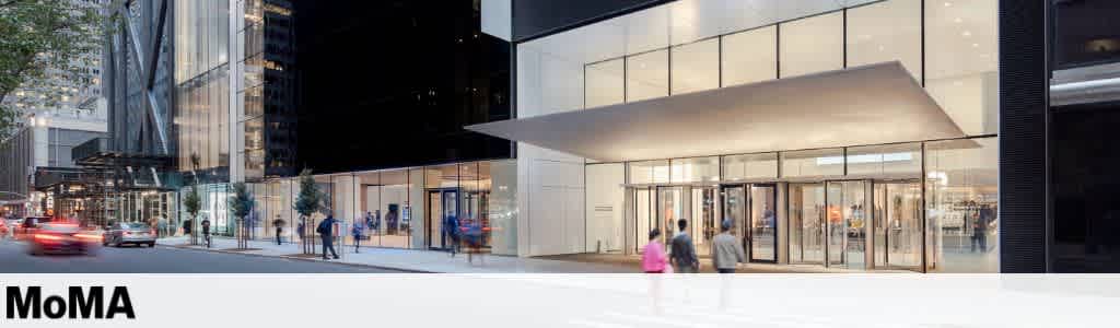 Street view of a busy museum entrance with glass facade and modern design. People walk along the sidewalk while cars drive by. A large canopy extends over the entrance, which is illuminated with warm lights, creating a welcoming atmosphere. Tall buildings surround the area, adding to the urban feel.