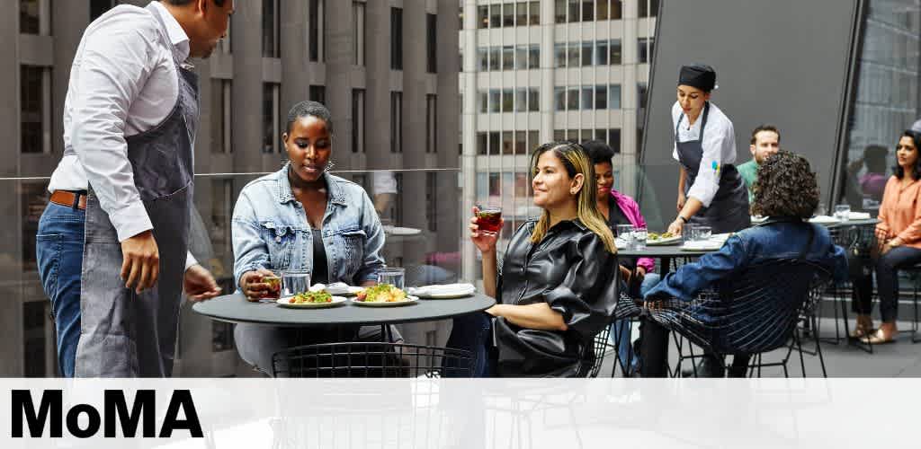 A group of people enjoy a meal on an outdoor terrace surrounded by city buildings. Individuals are seated at small, round tables, engaged in conversation. A waiter stands by, wearing an apron, while another server delivers a dish. Plates with colorful food and drinks are visible on the tables, creating a lively and social atmosphere.