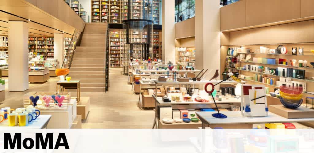 A spacious, well-lit store displays a variety of colorful products on tables and shelves, including mugs, books, and decorative items. A central staircase leads to an upper level lined with more merchandise. The store has a modern and vibrant atmosphere, with high ceilings and large windows.