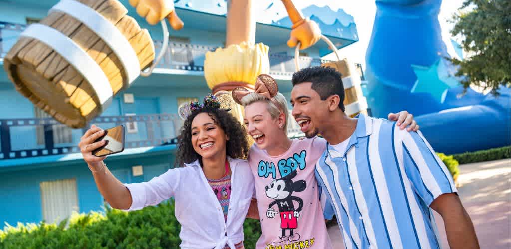 Three young adults smile and take a selfie together in front of a large Sorcerer Mickey statue and whimsical decorations at a theme park or resort.