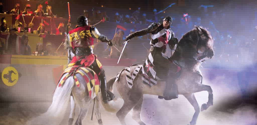 Two knights in colorful armor engage in a dramatic jousting match on horseback. The arena is filled with spectators watching from tiered seating. The scene is set in an indoor venue with bright, colorful banners and a misty atmosphere. One knight wears red and yellow, while the other has black and white. The action is intense as both knights wield swords, capturing the excitement of this medieval-themed event.