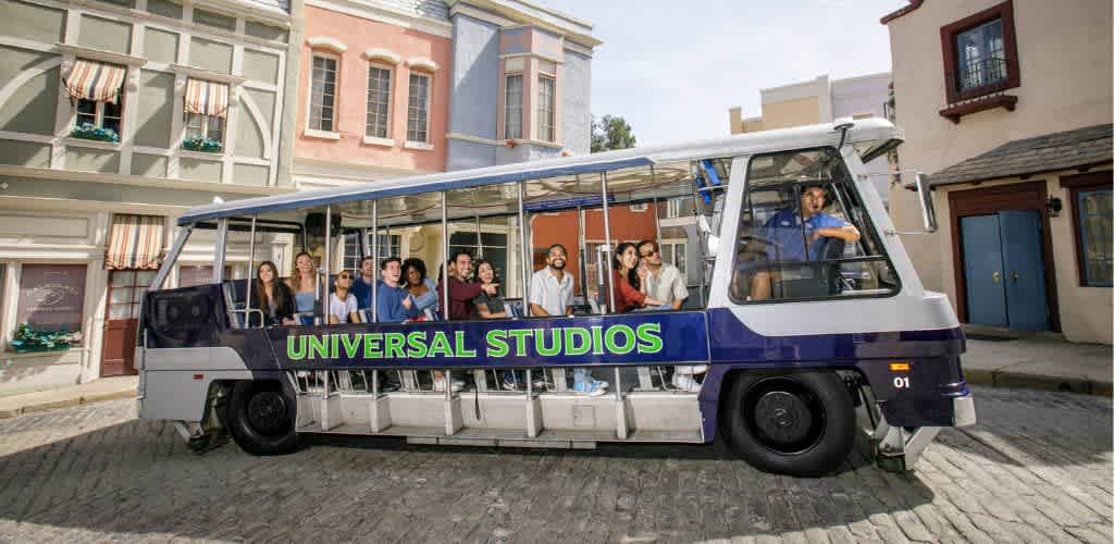 A group of people enjoy a tour on an open-air tram with Universal Studios printed on the side. The tram moves through a street with colorful buildings and striped awnings, capturing the charm of a lively movie set. The diverse group of passengers, along with the driver, are seated comfortably as they observe the surroundings, embraced by the vibrant and immersive experience. The scene represents entertainment and adventure, ideal for visitors seeking fun and exploration.