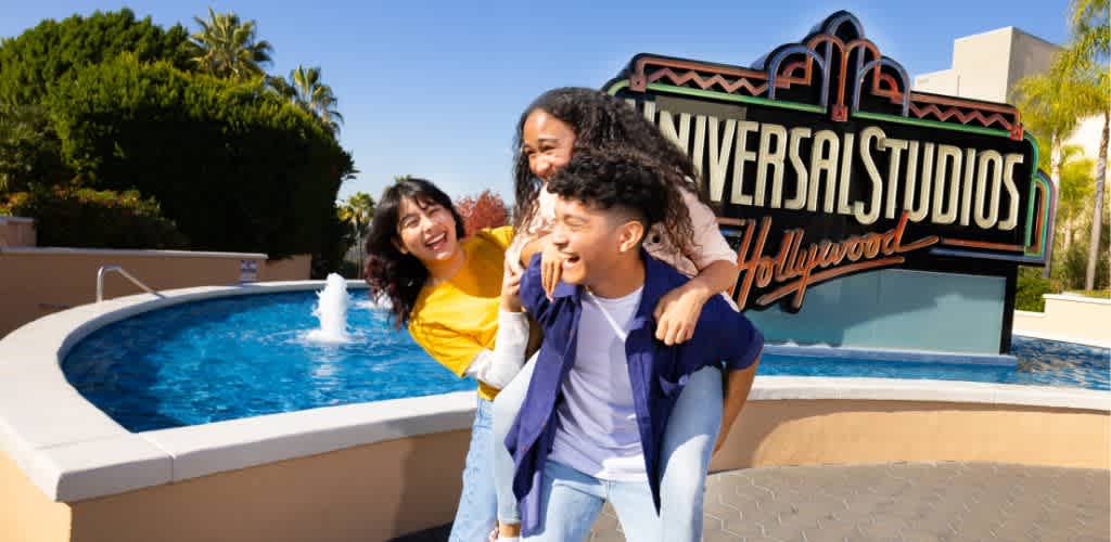 A joyful group of friends is seen at Universal Studios Hollywood. One person is giving a piggyback ride to another while a third person laughs beside them. They stand near a fountain with the iconic Universal Studios Hollywood sign in the background, surrounded by greenery and clear skies. The image captures a moment of fun and excitement, emphasizing the thrilling experiences available at the theme park.