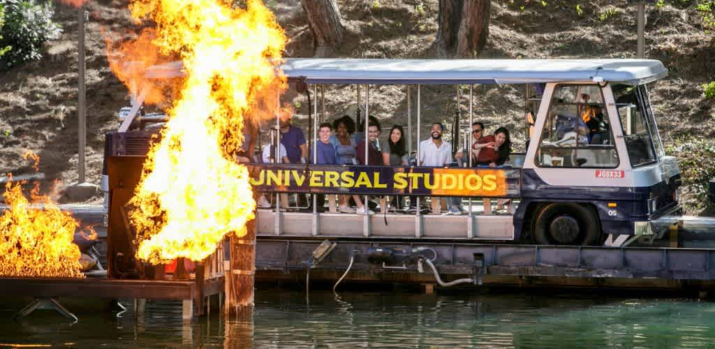 A tour tram at Universal Studios passes by a special effects display with large, controlled flames shooting up beside it. Passengers on the tram observe the scene, which adds excitement to the studio tour experience. The backdrop includes a hillside, enhancing the adventure setting.