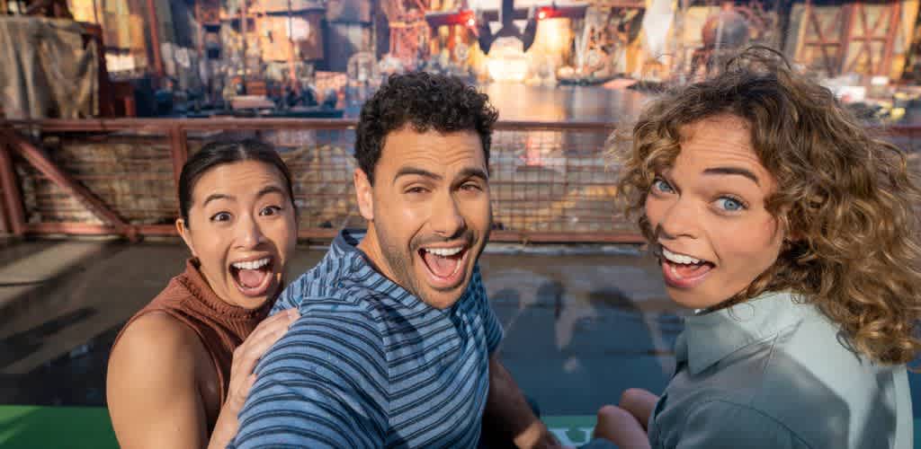 A diverse group of three people are joyfully posing for a selfie in an outdoor amusement park setting. They are seated with an array of colorful attractions and structures visible in the background, conveying an atmosphere of excitement and fun. The sunny weather adds to the lively and vibrant experience.