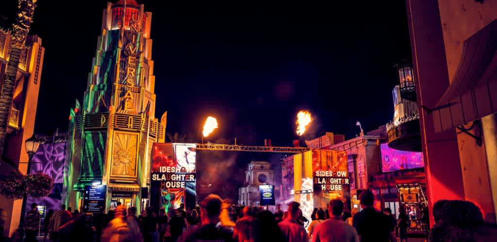 A lively theme park scene at night, vibrant lights illuminate the architecture and colorful signs. Visitors stroll past an art deco style tower with neon accents and colorful wall projections. Fiery torches are lit, adding to the excitement. The crowd enjoys the active nightlife atmosphere, surrounded by an array of attractions and entertainment options.