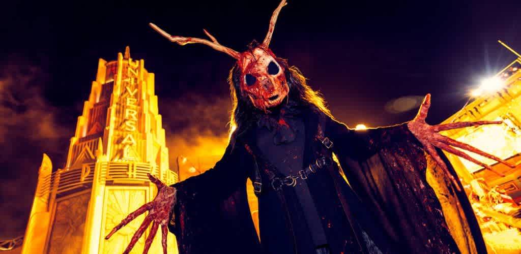 A person in a dark, creepy costume with a mask featuring antlers stands with arms outstretched, creating a spooky atmosphere. The background shows a brightly lit art-deco style building at night with the word Universal visible on signage. The scene is likely from a haunted event or attraction, capturing the thrill and mystery of Halloween festivities.