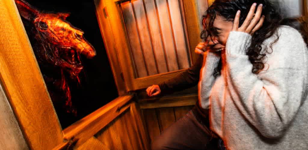 A person reacts in fright, covering their ears while standing in front of a wooden fence. On the other side of the fence, a menacing creature with a distorted, snarling face looms, creating a spooky and intense atmosphere. The lighting is warm, casting dramatic shadows and enhancing the eerie mood of the scene.