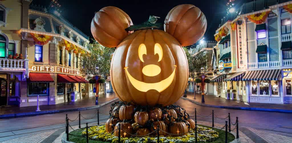 A giant pumpkin carved with a smiling face and large round ears resembling a famous character sits at the center of a festive street. The pumpkin is surrounded by smaller pumpkins and floral arrangements. Vibrant lights illuminate nearby shops adorned with fall decorations. This lively scene captures a whimsical and colorful atmosphere, suitable for a family-friendly environment.