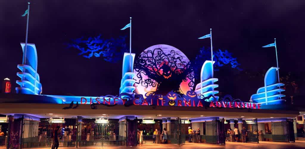 Nighttime view of an amusement park entrance adorned with Halloween decorations. A large, illuminated tree silhouette features spooky faces against a glowing round backdrop. Blue and purple lights highlight the art deco-style entrance, with flags perched atop tall pillars. People gather near the gate, creating a festive atmosphere.