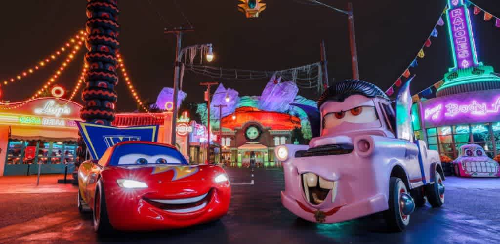 Colorful night scene featuring two animated cars with expressive faces posing in a lively, neon-lit town. The backdrop includes brightly illuminated buildings, strings of lights, and vibrant signage, creating a festive and whimsical atmosphere. The red car is smiling, while the tow truck has a playful expression. The setting evokes a sense of fun and adventure, perfect for fans of animated automotive characters.