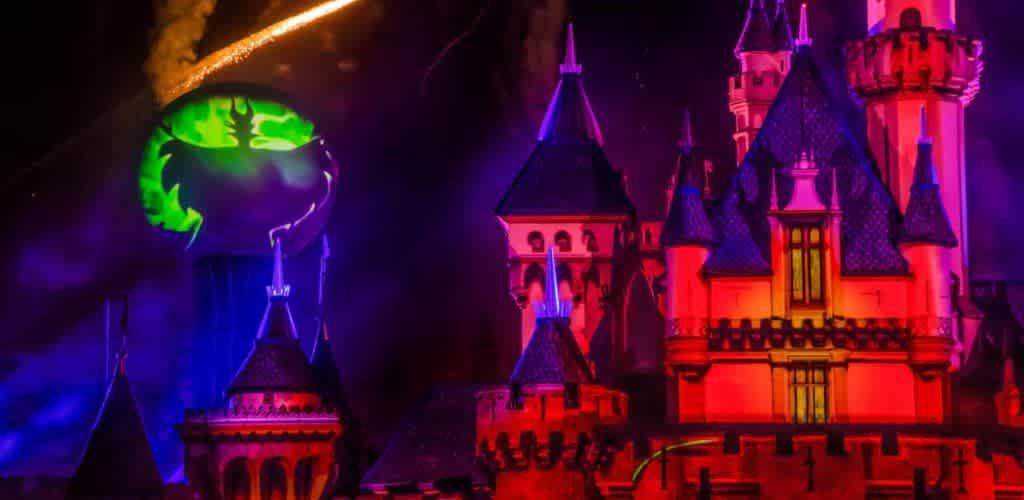 A castle is illuminated in vibrant pink and purple lights during a nighttime event. A large, menacing green face is projected on a circular screen to the left of the castle tower. The scene is dramatic, enhanced by a streak of golden fireworks in the dark sky above. The atmosphere is magical and slightly eerie, perfect for a themed celebration.