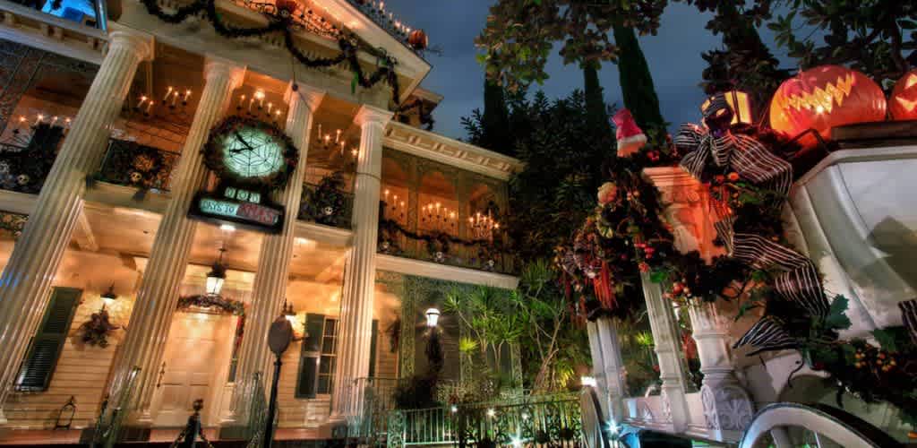 Festively decorated mansion with grand columns and warm lighting. A large clock dominates the facade, surrounded by garlands. The porch is adorned with glowing jack-o-lanterns and holiday wreaths. A spooky figure in a striped outfit and Santa hat stands nearby. Tall trees frame the scene against a dusky sky, creating a magical holiday atmosphere.