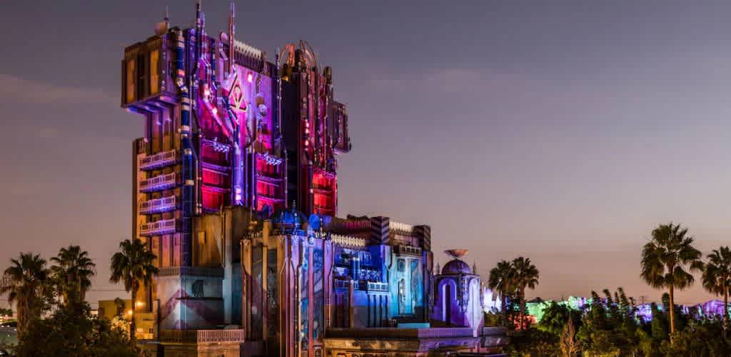 A colorful tower is illuminated at night with vibrant lights in shades of purple, blue, and red. The structure features distinctive architectural patterns and elements. Surrounding the tower are palm trees, with a clear evening sky in the background, adding to the dramatic and enchanting atmosphere of the scene.