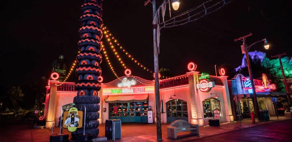 The image displays a vibrant night scene at a themed amusement park area. Neon lights in red, orange, and green illuminate a retro-style building, with signage that suggests a tire shop and cafe. A tall stack of stylized tires stands prominently in front, adorned with glowing neon lights. Strings of colorful bulbs stretch across the scene, adding to the lively ambiance. The setting evokes a sense of nostalgia and fun.