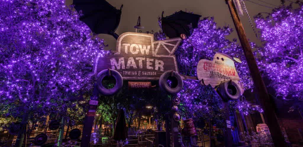 The image shows a whimsical setting with a colorful tow truck-themed sign featuring Tow Mater Towing and Salvage, suspended among trees adorned with vibrant purple lights. The atmosphere is playful and inviting, evoking a festive and magical outdoor experience.