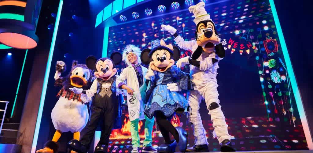 Characters in colorful costumes stand on a vibrant stage during a lively performance. The backdrop is filled with bright lights and festive decorations, creating an energetic and fun atmosphere. A whimsical setting perfect for a Halloween-themed event.