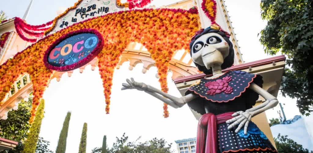 A colorful archway adorned with orange flowers welcomes visitors at Plaza de la Familia, featuring a vibrant sign for Coco. Below stands a large skeleton statue dressed in traditional Mexican attire, with an inviting gesture. The scene is set against a backdrop of trees and clear sky, celebrating Mexican culture in a festive atmosphere.