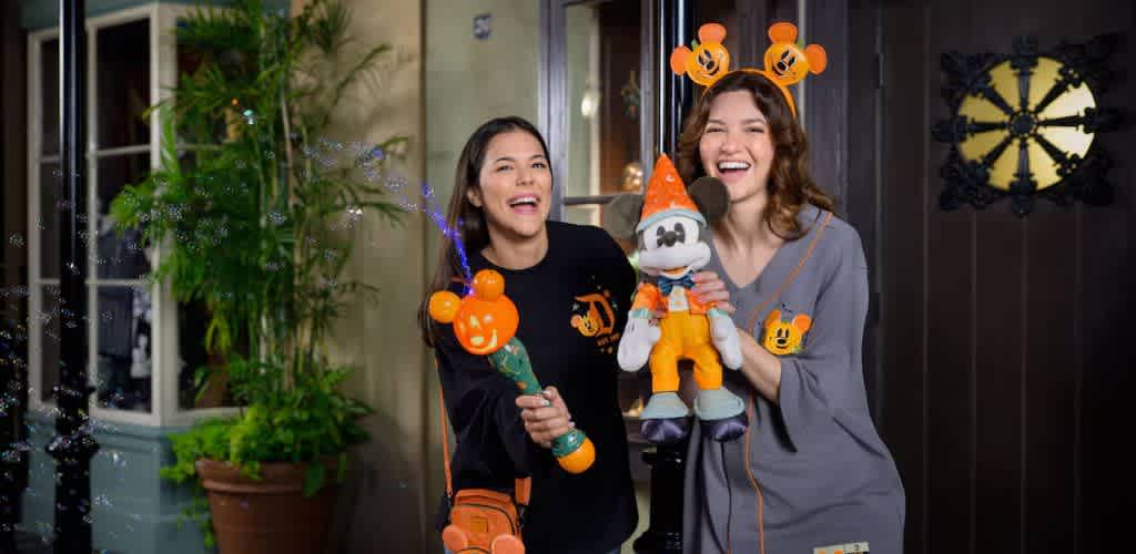 Two people smiling and enjoying a festive atmosphere. One holds a bubble wand shaped like a pumpkin, creating bubbles, while the other holds a plush toy wearing Halloween-themed attire. Both wear headbands with themed decorations. The background features a potted plant and a decorative wall element, enhancing the cheerful scene.