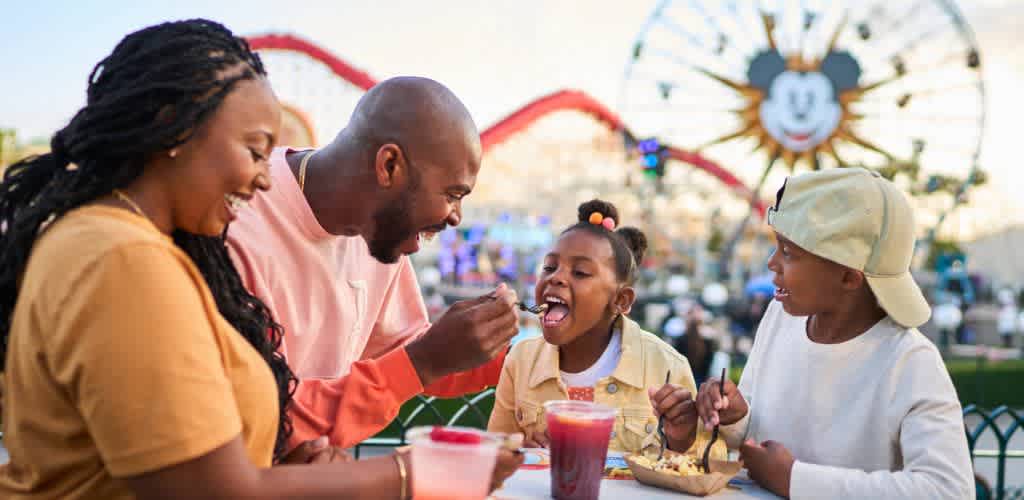 A family enjoys a day at an amusement park, sitting and sharing snacks at a table. In the background, a Ferris wheel with a smiling face and colorful attractions are visible. The atmosphere is bright and cheerful, reflecting a joyful outing for all.